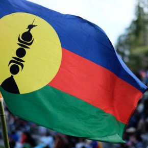 drapeau de la kanaky en premier plan, en second plan flou un rassemblement de personnes et plusieurs autres drapeaux kanaks