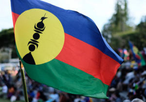 drapeau de la kanaky en premier plan, en second plan flou un rassemblement de personnes et plusieurs autres drapeaux kanaks